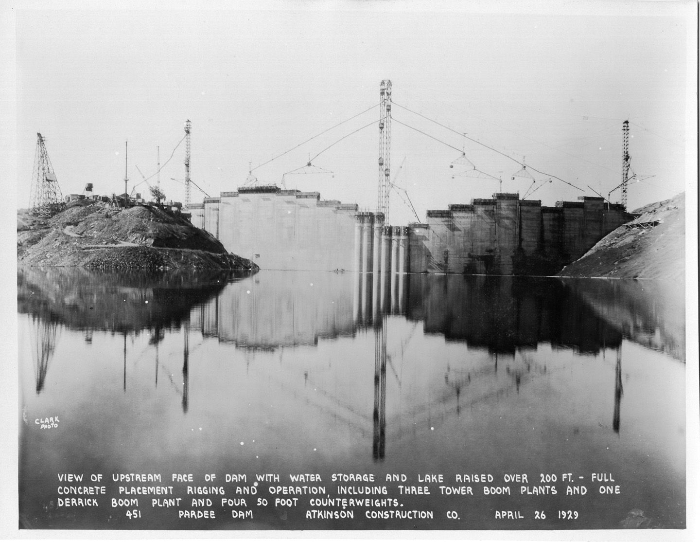 View of upstream face of dam with water storage and lake raised over 200 ft. - full concrete placement rigging and operation including three tower boom plants and one derrick boom plant and four 50 foot counterweights.(April 1929)	