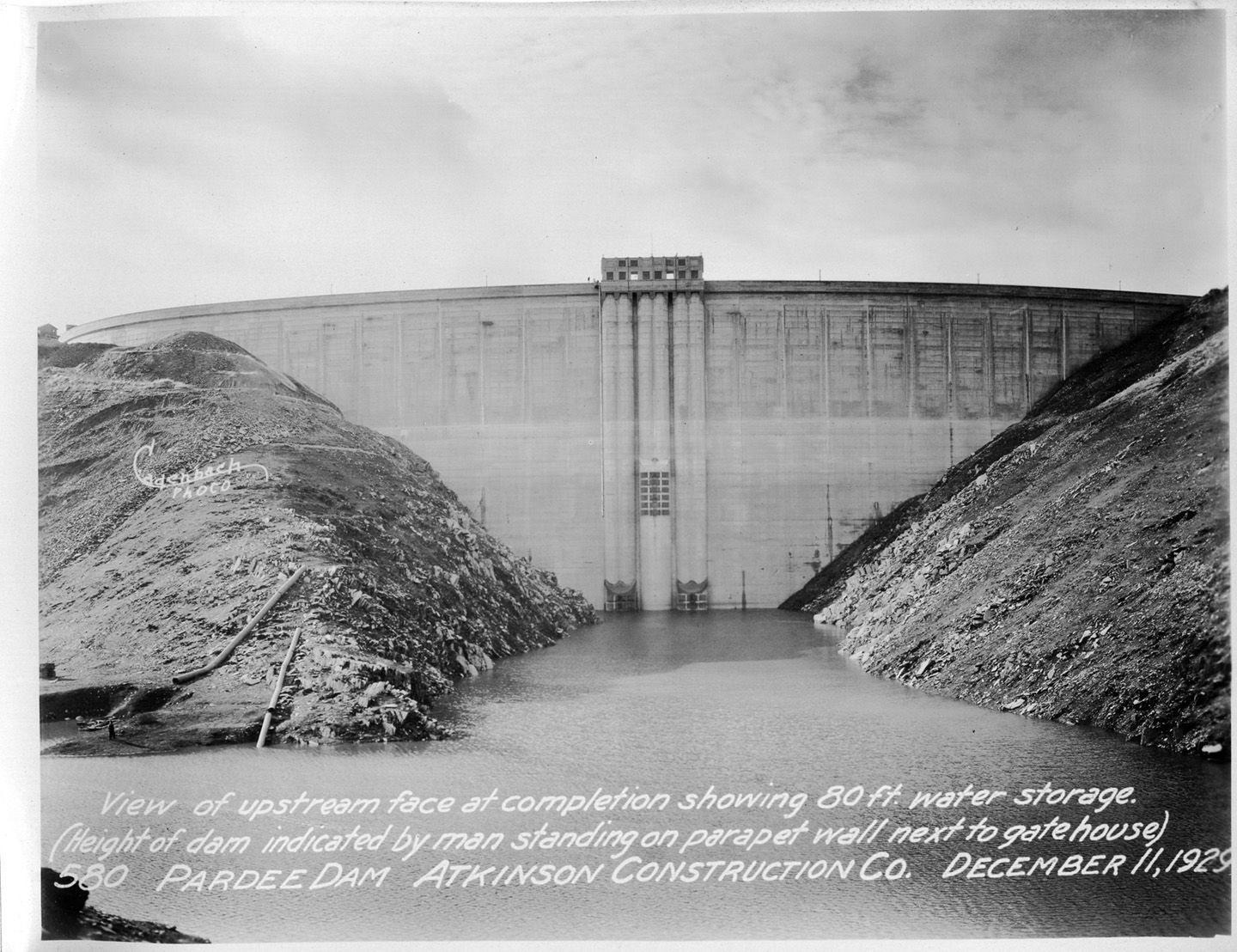 View of upstream face at completion showing 80 ft. water storage. (Height of dam indicated by man standing on parpet wall next to gatehouse). (December 1929)	