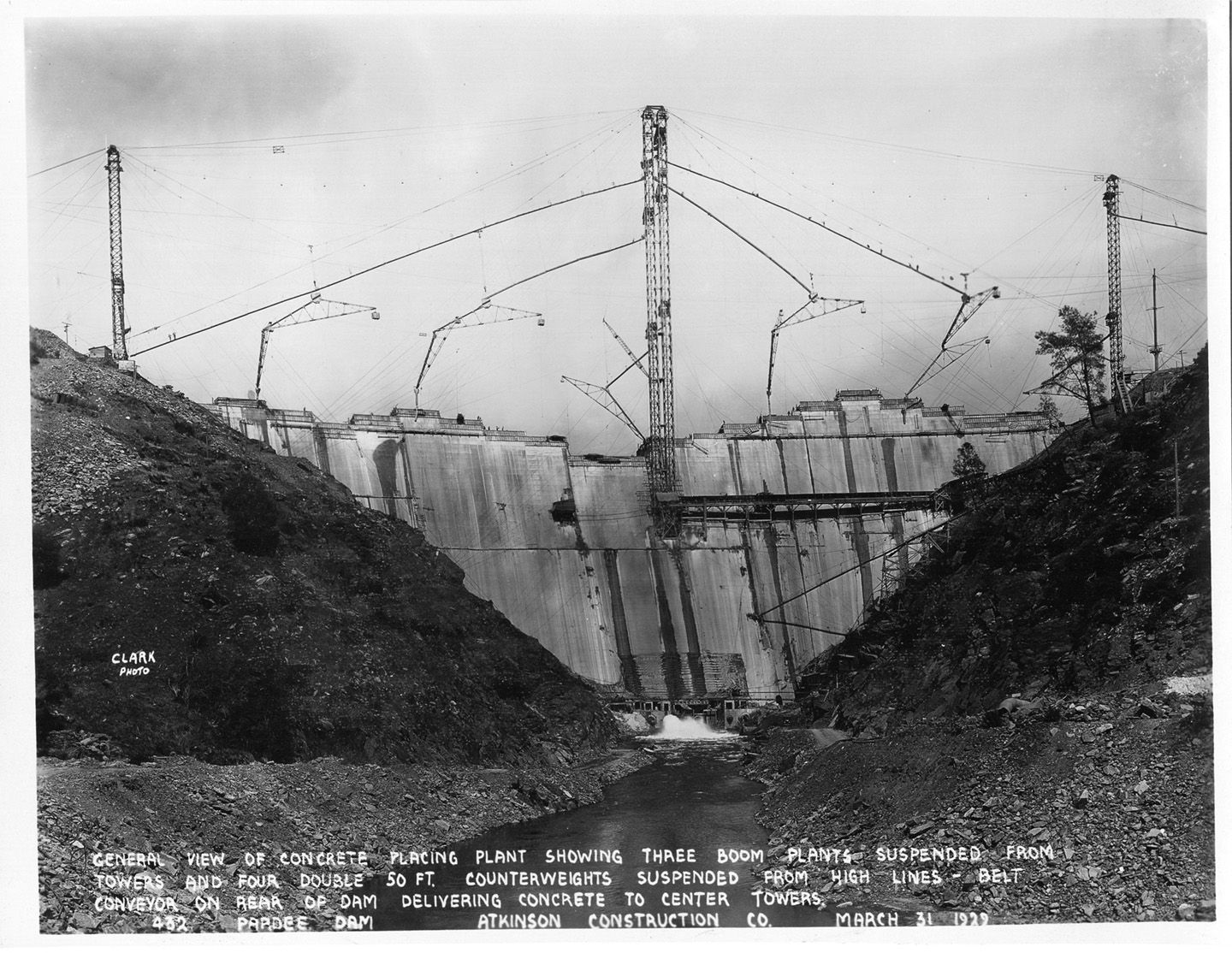 General view of concrete placing plant showing three boom plants suspended from towers and four double 50 ft. counterweights suspended from high lines - belt conveyor on rear of dam delivering concrete to center towers. (March 1929)	