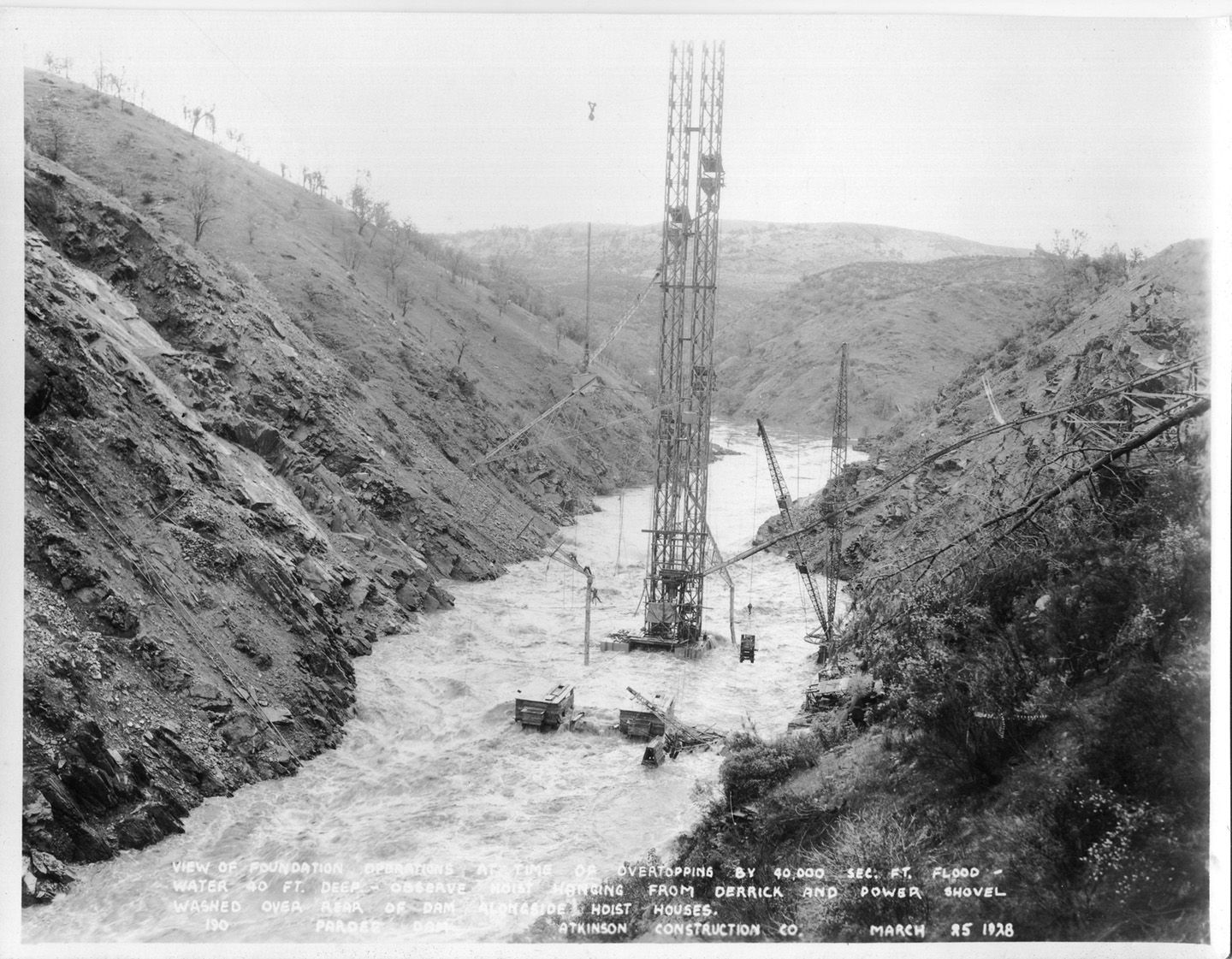 View of foundation operations at time of overtopping by 40,000 sec. ft. flood - water 40 ft. deep - observe host hanging from derrick and power shovel washed over rear of dam alongside hoist houses. (March 1928)