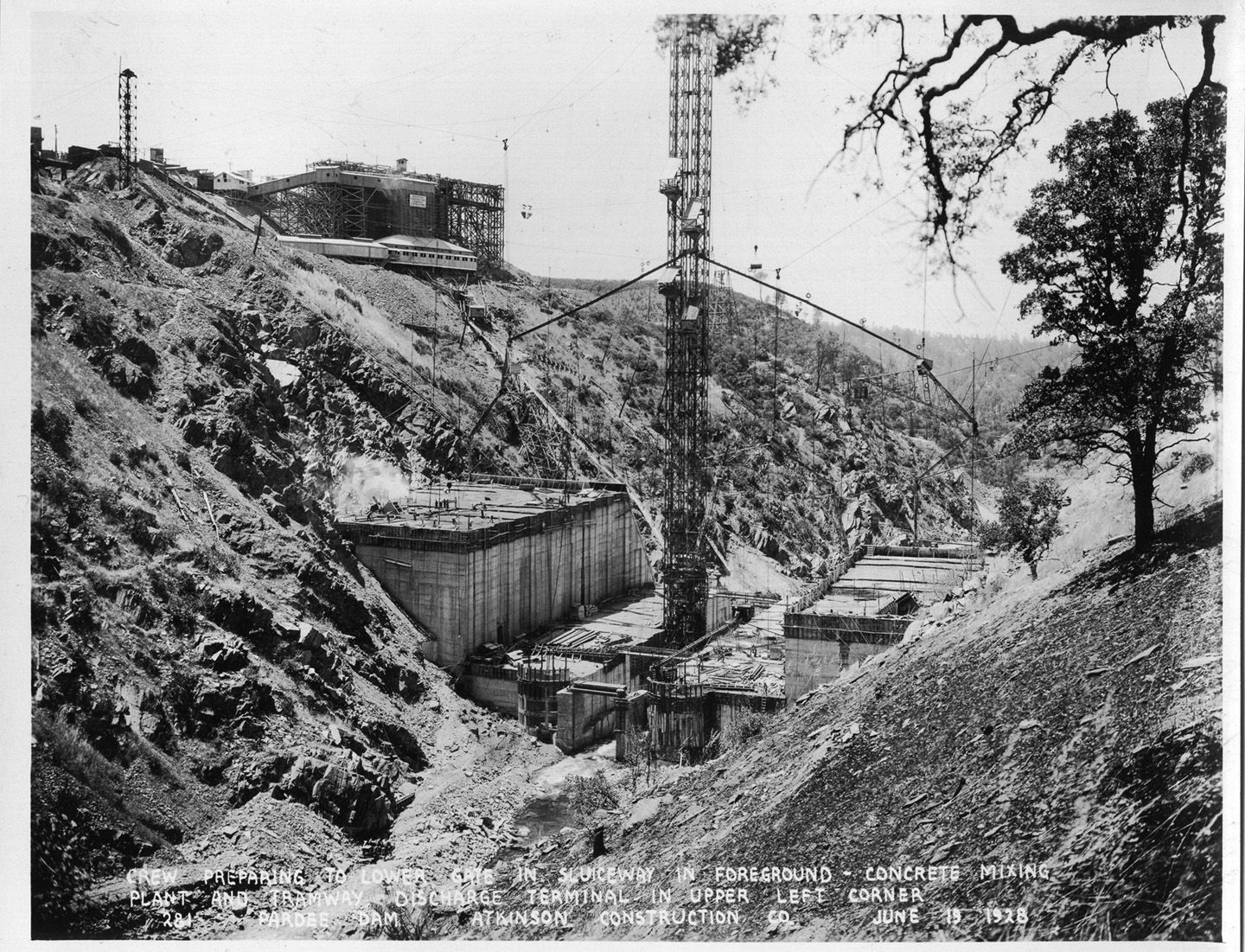 Crew preparing to lower gate in sluiceway in foreground - concrete mixing plant and tramway discharge terminal in upper left corner. (June 1928)